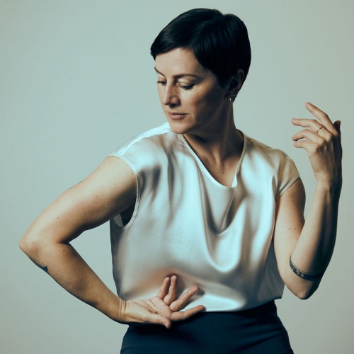 portrait of helene simoneau in colder muted tones. helene is in a dancer's post with right hand supinated and pressed to her waist while the left is bent upwards facing her. helene is looking down and to the side and has short black hair and is wearing a silken white blouse